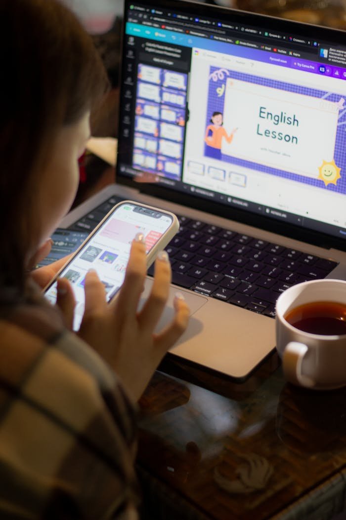 Adult woman multitasking, using laptop and smartphone while studying English with tea on a table.